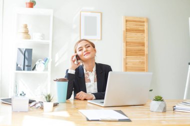 Smiling businesswoman in suit using laptop while talking on mobile phone at desk in home office