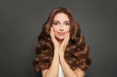 Pretty young woman with natural makeup, clean skin and long healthy curly brown hair smiling on gray background.