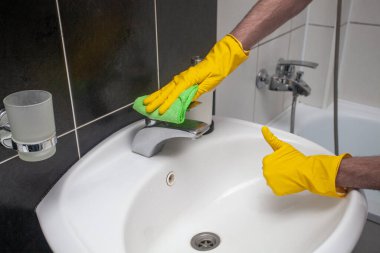 Hands with cloth cleaning white sink and faucet and showing thumb up