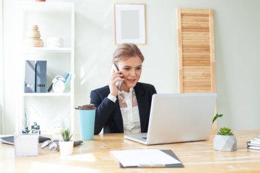 Portrait of young business woman sitting in front of her laptop and talking on mobile phone