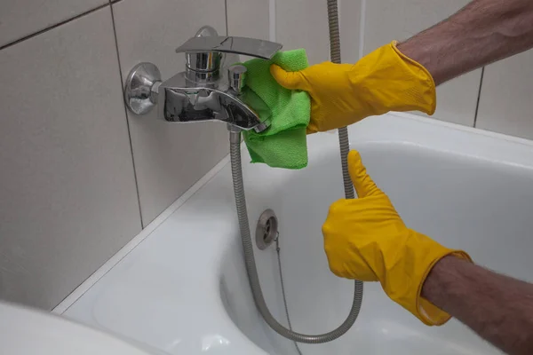 Hands with rag and detergent spray cleaning a faucet in the bathroom