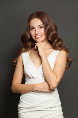 Cute woman in white dress smiling and standing with crossed arms on gray background.