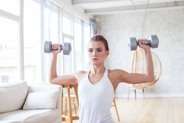 Athletic woman in sportswear doing fitness exercises with dumbbells at home. Sport and recreation concept.