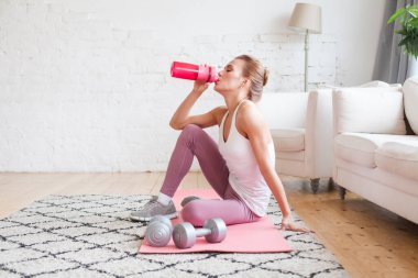 Young woman drinking water or proteine shake after fitness exercises at home interior 