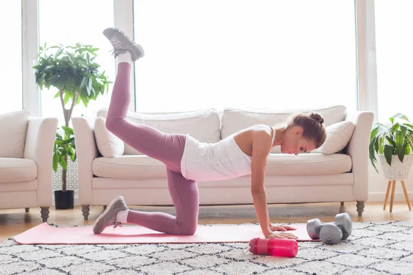 Sport and recreation concept. Young woman doing donkey kicks exercise indoor at home interior.