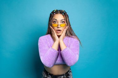 Güzel, oldukça şaşırmış, sarı gözlüklü, yanaklarıyla el ele tutuşan, heyecanlı bir kadın. Mavi arka planda güzel haberlerin tadını çıkarıyor.