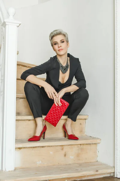 Fashion style portrait of perfect blonde woman in stylish suit with red handbag posing indoor