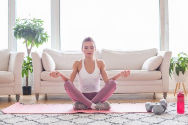 Genç, formda bir kadın evde meditasyon yapıyor yoga minderinde oturuyor ve gözleri kapalı gülümsüyor. Farkındalık meditasyon konsepti, Sağlıklı Yaşam tarzı
