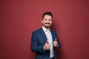 Mutlu, takım elbiseli bir işadamı kırmızı stüdyo duvarına başparmak işareti yapıyor.