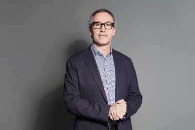Middle-aged man wearing glasses standing with folded arms over a grey background looking at camera