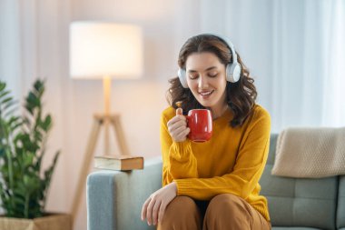Neşeli genç bir kadın kulaklıkla müzik dinliyor evdeki kanepede oturuyor..