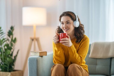 Neşeli genç bir kadın kulaklıkla müzik dinliyor evdeki kanepede oturuyor..