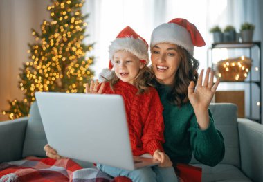 Anne ve kızı, Noel için dekore edilmiş oturma odasında dizüstü bilgisayar kullanan biriyle sohbet ediyorlar..