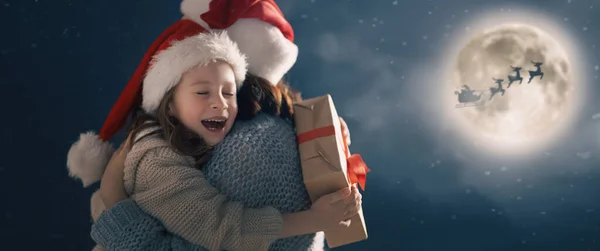 圣诞快乐 节日快乐 可爱的小孩和妈妈在一起 圣诞老人骑雪橇在月亮的天空中飞舞 在黑暗背景下享受假期的家庭 — 图库照片