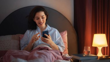 Mutlu, sıradan, güzel bir kadın evdeki yatakta telefon kullanıyor..