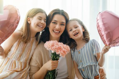 Mutlu bir gün! Çocuklar annelerini kutluyor ve ona çiçek veriyorlar. Anne ve kızlar gülümsüyor ve sarılıyorlar. Aile tatili ve birliktelik.