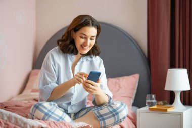 Mutlu, sıradan, güzel bir kadın evde bir yatağın üzerinde oturan telefonu kullanıyor..