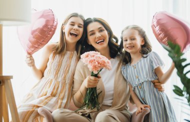 Mutlu bir gün! Çocuklar annelerini kutluyor ve ona çiçek veriyorlar. Anne ve kızlar gülümsüyor ve sarılıyorlar. Aile tatili ve birliktelik.