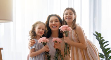 Mutlu bir gün! Çocuklar annelerini kutluyor ve ona çiçek veriyorlar. Anne ve kızlar gülümsüyor ve sarılıyorlar. Aile tatili ve birliktelik.