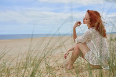 Gülümseyen, hayat dolu genç bir kadın kumsalda dinleniyor, kumullarda oturuyor, denize bakıyor, güneşin tadını çıkarıyor.