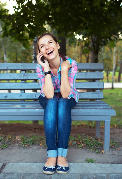A portrait of a smiling woman in a park on a bench talking on th — Stock Photo, Image