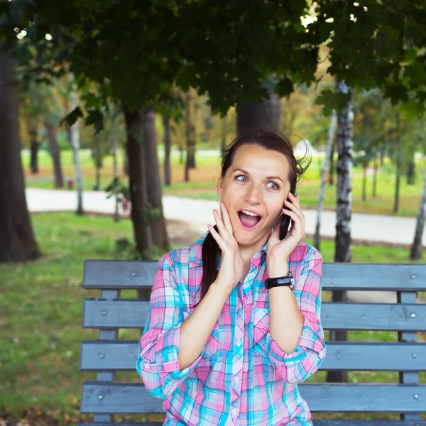 A portrait of a smiling woman in a park on a bench talking on th — Stock Photo, Image