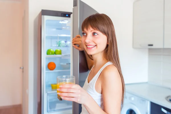 Giovane ragazza bruna con un bicchiere di succo vicino al frigorifero — Foto Stock