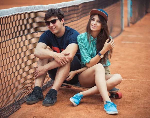 Couple sitting on a skateboard on the tennis court — Stock Photo, Image