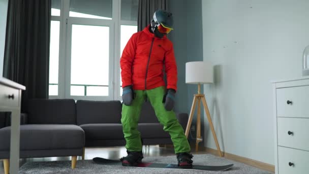 Śmieszne nagranie. Mężczyzna przebrany za snowboardzistę przedstawia snowboard na dywanie w przytulnym pokoju. Czekam na śnieżną zimę. Zwolniony ruch — Wideo stockowe