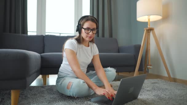 Casualmente mujer vestida con auriculares está sentado en la alfombra con el ordenador portátil y trabajando en una habitación acogedora. Trabajo remoto fuera de la oficina. — Vídeos de Stock