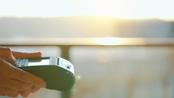 クローズアップ、女性は銀行ターミナルで注文を支払うためにスマートフォンのキャッシュレス財布NFC技術を使用しています。太陽の光を背景に男性の手でカードマシン — ストック動画