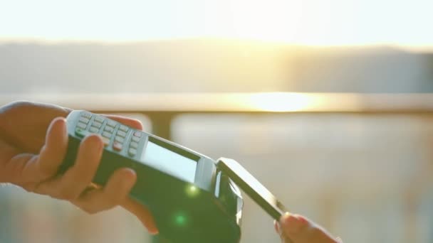 Close-up, woman using smartphone cashless wallet NFC technology to pay order on bank terminal. Card machine in male hand on sunlight background — Stock Video