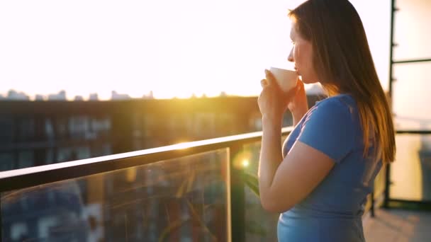 Mulher com uma xícara de café em pé na varanda e admirar o pôr do sol. — Vídeo de Stock