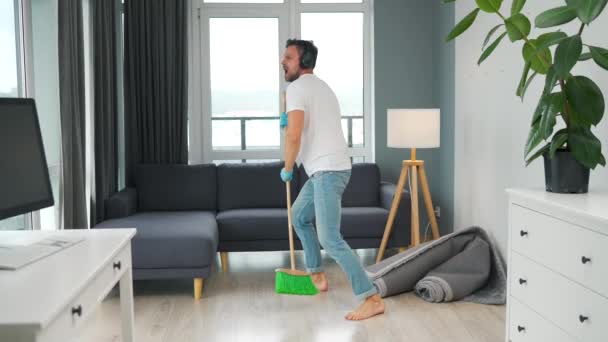 Hombre con auriculares limpiando la casa y divirtiéndose bailando y cantando con una escoba. Movimiento lento — Vídeo de stock