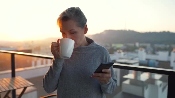 Vrouw begint haar dag met een kopje thee of koffie en het controleren van e-mails in haar smartphone op het balkon bij dageraad. — Stockvideo