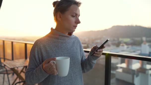 Woman starts her day with a cup of tea or coffee and checking emails in her smartphone on the balcony at dawn, slow motion. — Stock Video