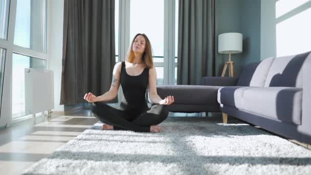 Junge kaukasische Frau im schwarzen Overall sitzt in Lotusposition und macht Yoga zu Hause, um Flexibilität und Gleichgewicht zu entwickeln — Stockvideo