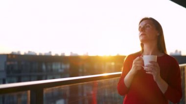 Elinde bir fincan kahve olan kadın balkonda duruyor ve gün batımını seyrediyor. Yavaş çekim