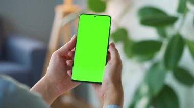 Evde dikey modda yeşil ekranlı akıllı telefon kullanan bir kadın var. Kız internette geziniyor, içeriği izliyor, videolar izliyor. POV.