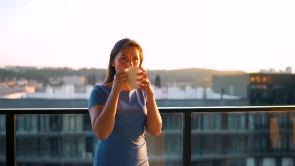 Elinde bir fincan kahve olan kadın balkonda duruyor ve gün batımını seyrediyor. Yavaş çekim — Stok video