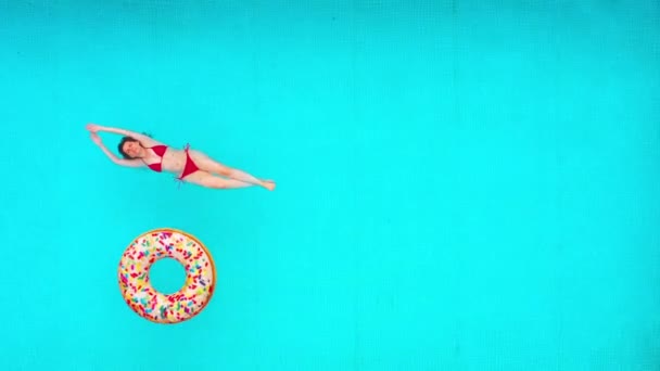 从上往下看，就像一个穿着红色泳衣的女人躺在游泳池里。多色的充气甜甜圈飘浮在她身边。放松的概念 — 图库视频影像