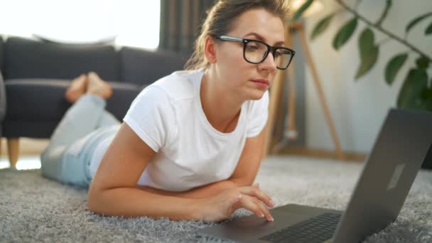 Жінка в окулярах лежить на підлозі і працює на ноутбуці. Концепція віддаленої роботи . — стокове відео