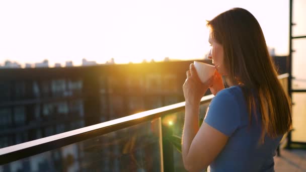 Frau mit einer Tasse Kaffee steht auf dem Balkon und bewundert den Sonnenuntergang. Zeitlupe — Stockvideo