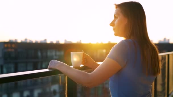 Frau mit einer Tasse Kaffee steht auf dem Balkon und bewundert den Sonnenuntergang. — Stockvideo