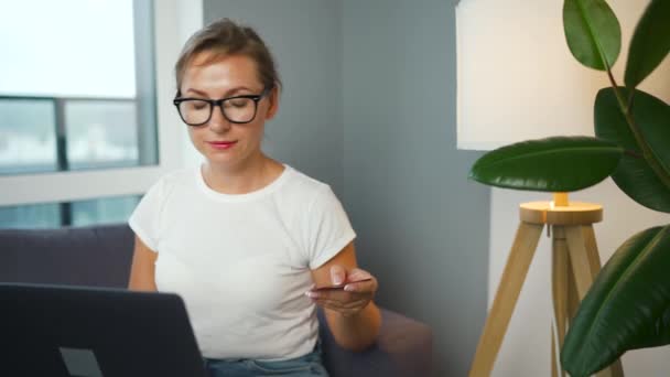 Mujer con gafas se sienta en un sofá en una habitación acogedora y hace una compra en línea con una tarjeta de crédito y un teléfono inteligente. Compras en línea, tecnología de estilo de vida — Vídeos de Stock