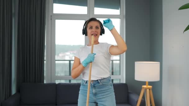 Frau mit Kopfhörer putzt das Haus und hat Spaß beim Singen mit dem Besen wie ein Star bei einem Konzert. Zeitlupe — Stockvideo