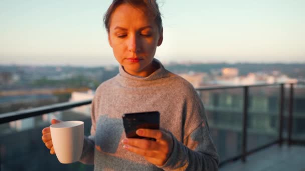 Vrouw begint haar dag met een kopje thee of koffie en het controleren van e-mails in haar smartphone op het balkon bij dageraad, slow motion. — Stockvideo