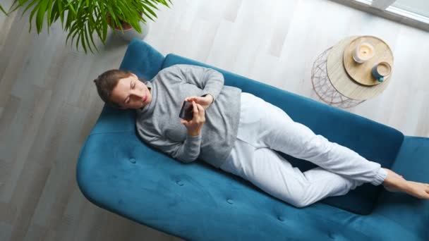 Vue aérienne d'une femme heureuse et détendue tenant un téléphone intelligent, utilisant des applications mobiles, regardant des vidéos drôles, s'amusant à bavarder dans les médias sociaux, couché sur le canapé à la maison. Caméra tourne dans le sens des aiguilles d'une montre Clip Vidéo