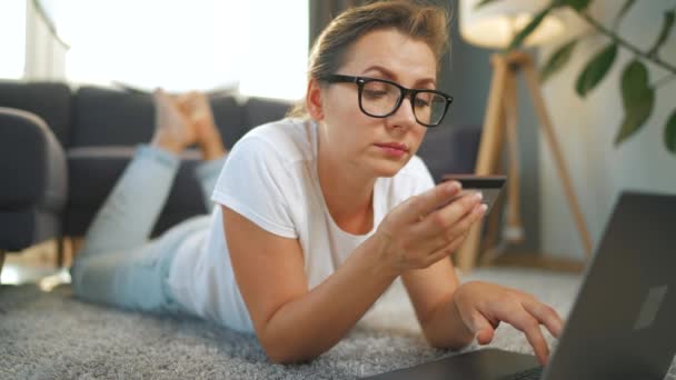 Kobieta w okularach leży na podłodze i dokonuje zakupu online za pomocą karty kredytowej i smartfona. Zakupy online, technologia stylu życia — Wideo stockowe