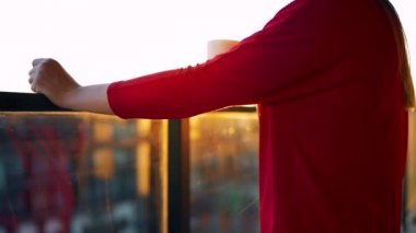 Kırmızı elbiseli bir kadın elinde bir fincan kahveyle balkonda duruyor ve gün batımını seyrediyor. Yavaş çekim